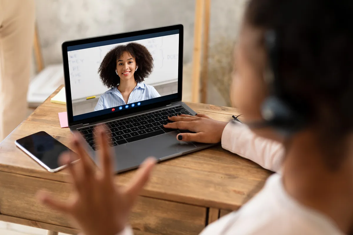 Learner on a video call with a smiling teacher on a laptop at a wooden desk