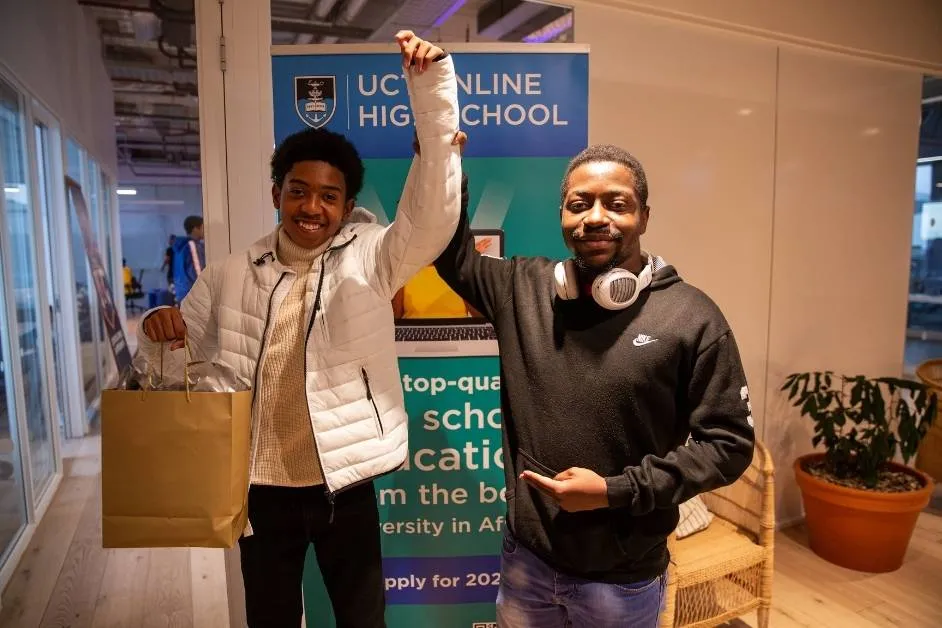 Two young men pose in front of a UCT Online High School banner, one raising the other’s arm in victory