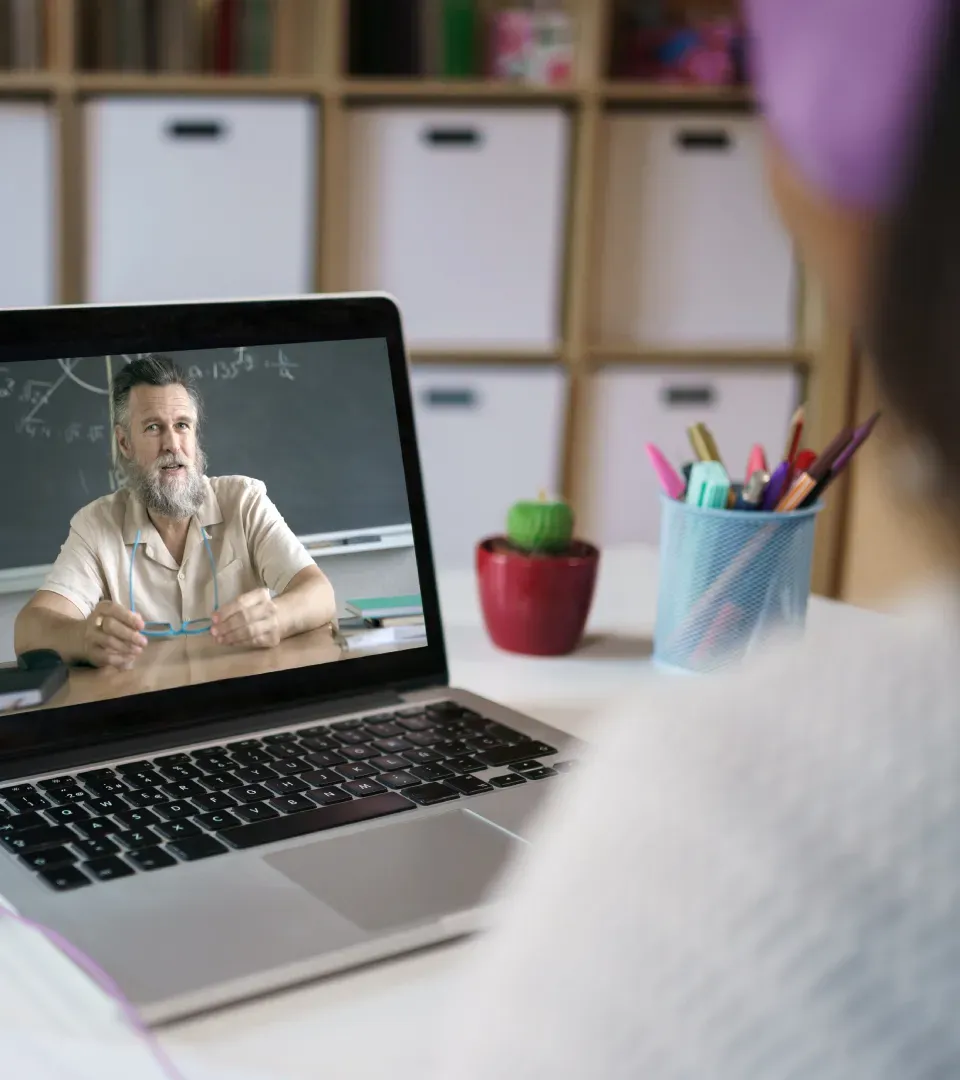 A learner watching a facilitator on a laptop, with a pot of coloured pens on the desk