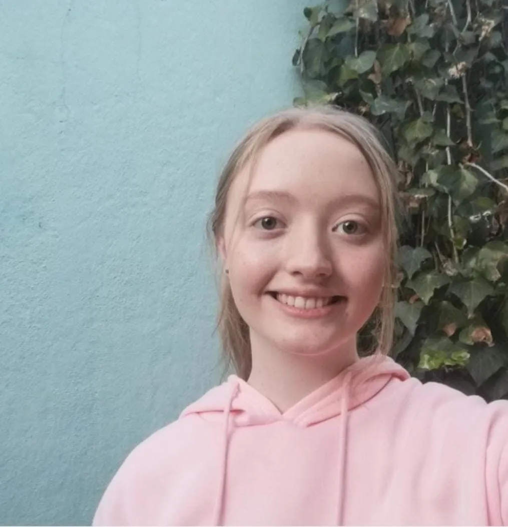 Portrait of Tanya du Toit, smiling in a pink hoodie in front of a blue wall and ivy